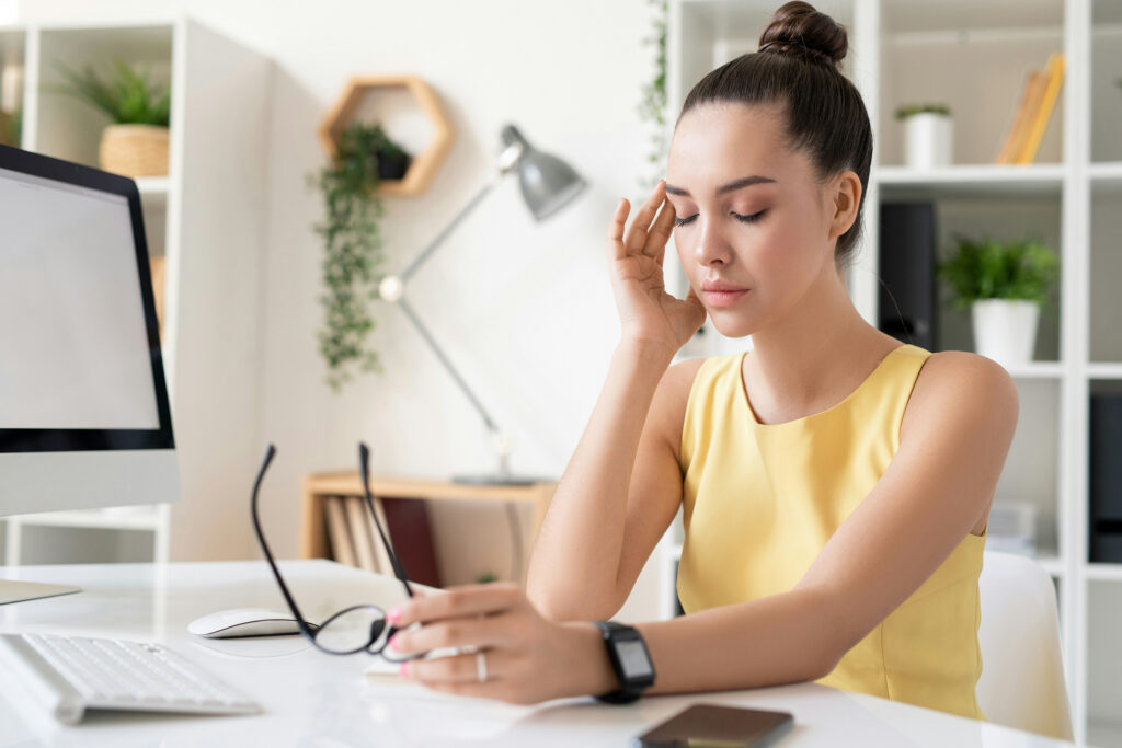 Woman sitting at desk with headache, scheduling chiropractic adjustment for natural pain relief