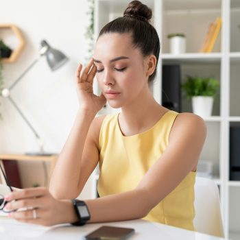 Woman sitting at desk with headache, scheduling chiropractic adjustment for natural pain relief